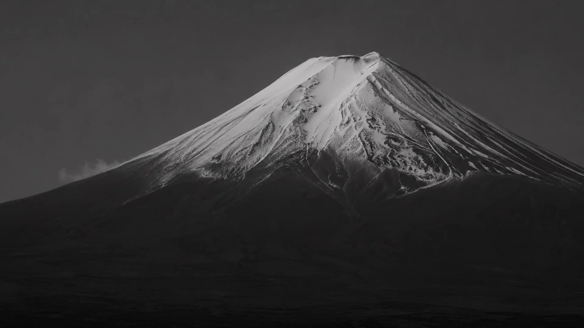 冠雪した富士山のモノクロ写真。暗い空を背景に、山頂の雪が白く浮かび上がっている
