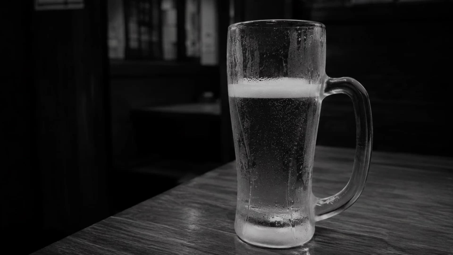 薄暗い居酒屋のテーブルに置かれた、水滴のついたビールジョッキ。モノクロ写真。